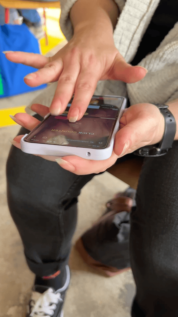 Rebecca's using two fingers on one hand on a smartphone to extremely rapidly press on a click counter app, then changes strategy and moves the phone to her lap and presses even faster