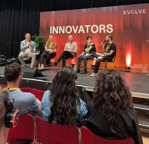 The panellists are seated on stage at Brighton Dome with a bright red backdrop with the word, Innovators written on it. Chair and panellists left to right: Ed Chinn, Catrina Baker-Bassett, Neil Clark, Fiona MacNeill, Mark Buss.